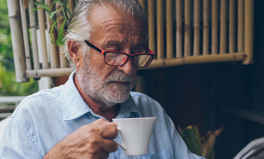 A silver haired man wearing glasses enjoys a mug of coffee while reading the newspaper.
